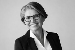 Smiling woman in a blazer with glasses, head-and-shoulders portrait in grayscale, neutral background.