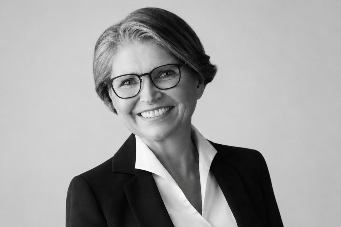 Smiling woman in a blazer with glasses, head-and-shoulders portrait in grayscale, neutral background.