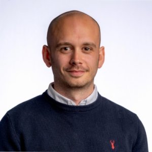 Head-and-shoulders portrait of a bald man in a navy knit sweater over a white collared shirt, small red emblem on the sweater, smiling softly against a light background.