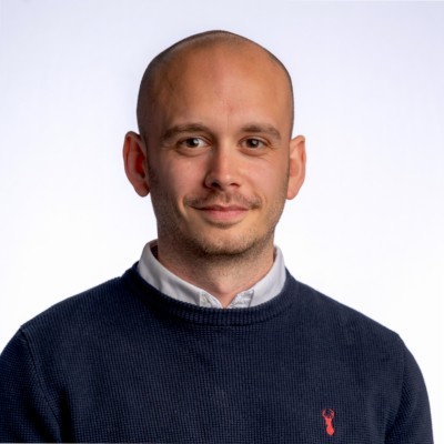 1656619590463 Head-and-shoulders portrait of a bald man in a navy knit sweater over a white collared shirt, small red emblem on the sweater, smiling softly against a light background.