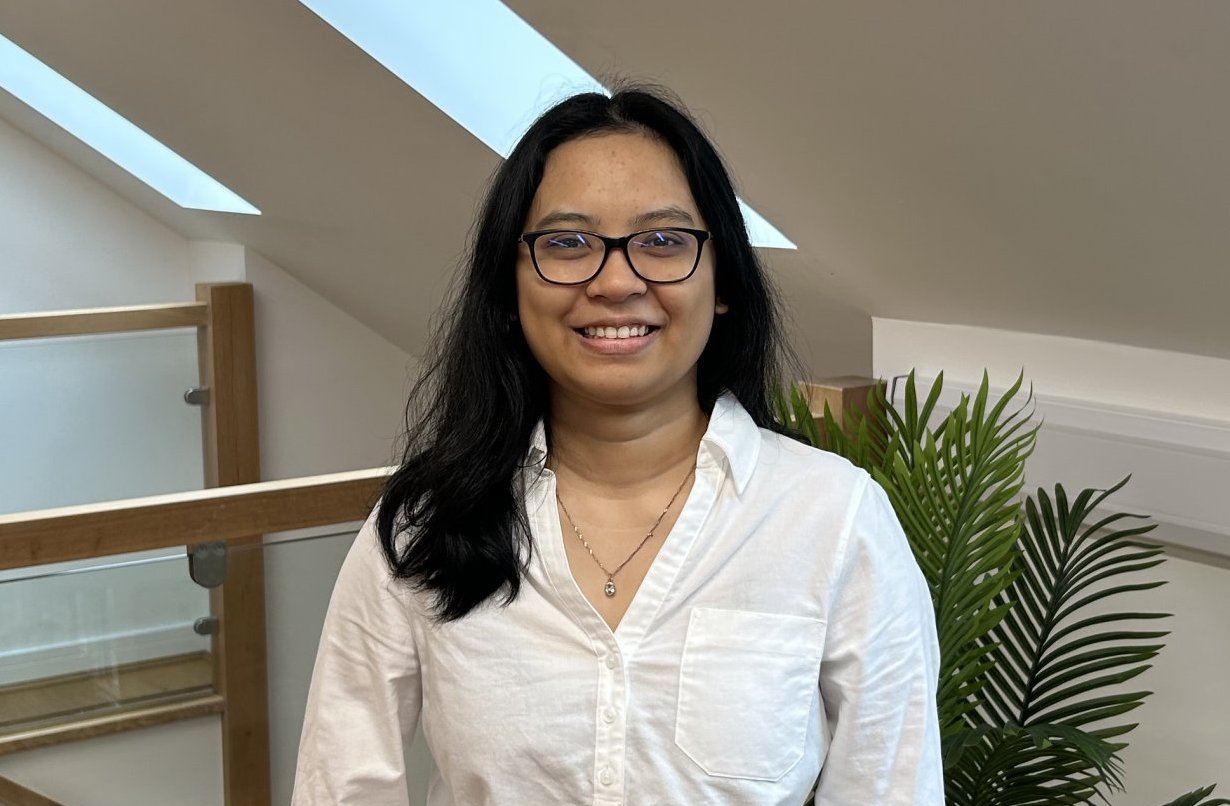 Smiling woman in a white shirt standing in a bright modern atrium with glass railing and a red tufted bench.