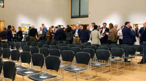 Group of professionals in business attire mingling and chatting in a conference room with rows of black chairs and light wood floors.