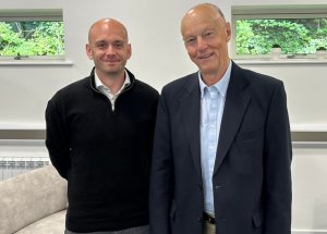 Two men posing indoors for a photo: one in a black sweater, the other in a blue shirt and dark blazer, both smiling.