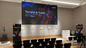 Stage setup for Torque & Truth event with a large central screen displaying the title and branding banners on either side, an arrangement of framed artwork along the back wall, and rows of black chairs facing the stage.