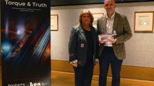 Two adults pose indoors beside a 'Torque & Truth' banner; woman in a dark blazer with patches and man in a plaid blazer holding a clear model airplane trophy.