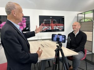 Two men in an office interview setup: one standing and gesturing, the other seated and smiling as a phone on a tripod records them.