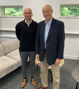 Two men stand side by side indoors, smiling, in a modern office with a beige sofa and windows behind them.
