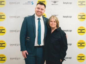 Smiling man in a teal suit and white shirt with a blue tie poses with a woman in a black jacket at a sponsor backdrop of Drivers of Change and Bentley buildings logos.